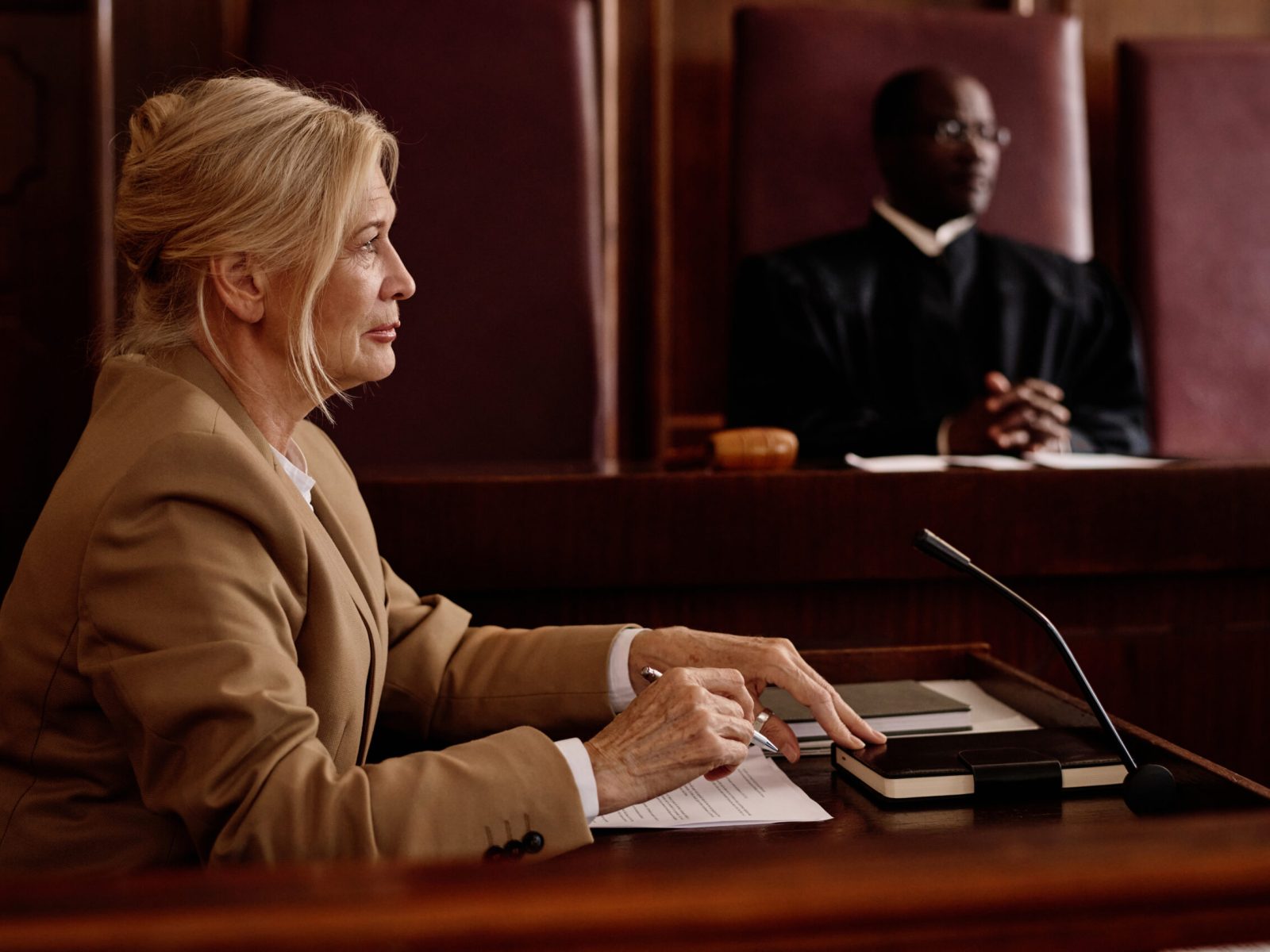 Side view of mature blond female witness or lawyer speaking in microphone while sitting against African American male judge